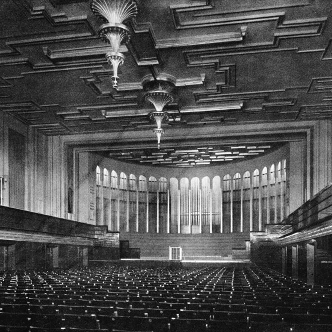 Schwarz-Weiß-Bild: Sitzränge vor einer großen Bühne in einem Saal mit Emporen rechts und links und hohen Decken, von den Decken hängen zwei Kornleuchter