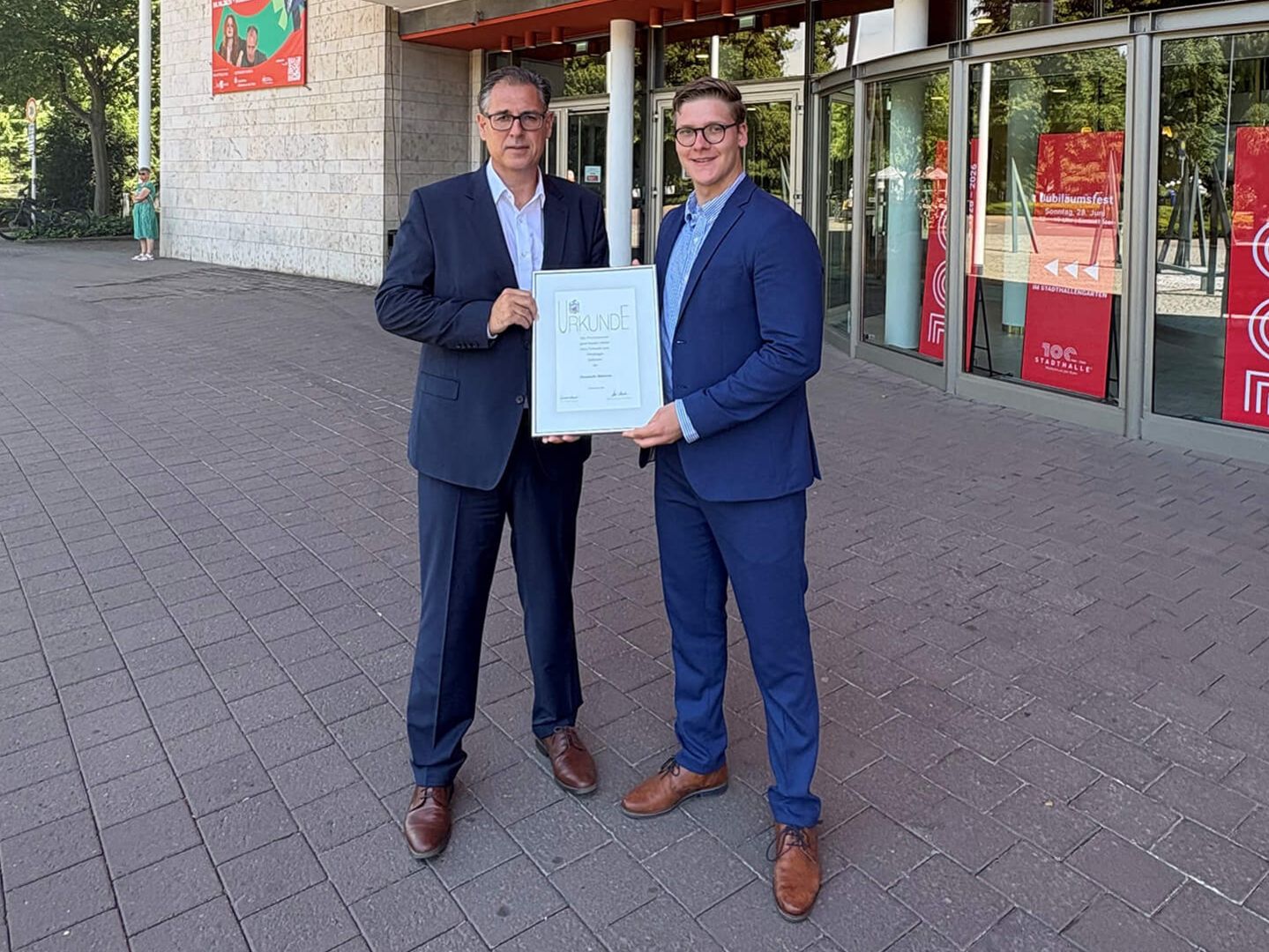 Niklas Mattheis (Stauder) und Michael Birr (MST) stehen vor dem festlich geschmückten Haupteingang der Stadthalle Mülheim und halten lächelnd eine Urkunde in die Kamera. 