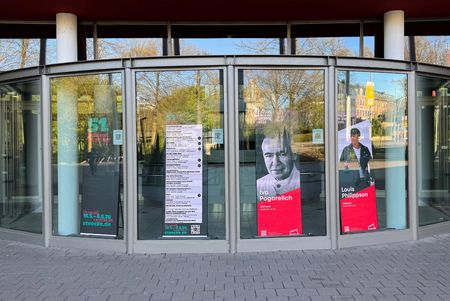 Vier hohe Bildschirme mit digitalen Veranstaltungsplakaten hinter großen Fensterscheiben am Haupteingang der Stadthalle Mülheim an der Ruhr